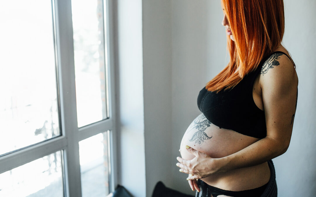 Mujer embarazada con tatuajes visibles observando por la ventana, en un momento de reflexión
