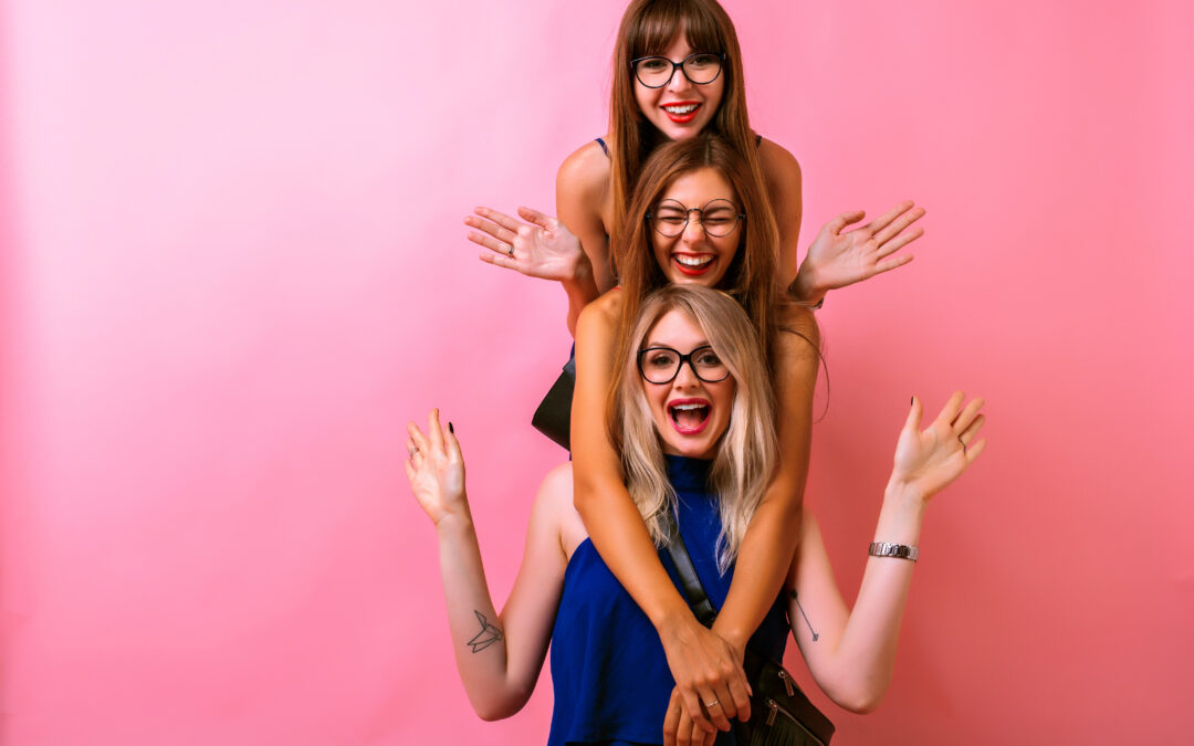 Tres mujeres jóvenes sonrientes posan juntas sobre un fondo rosado, transmitiendo alegría, confianza y bienestar femenino.